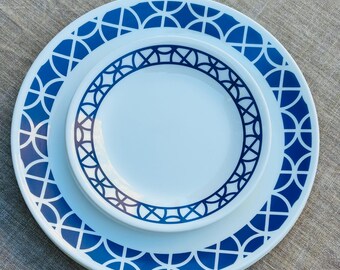 Puede incluir: Dos platos de cerámica blanca con un patrón geométrico azul. El plato más grande tiene un diseño repetitivo de arcos y diamantes azules alrededor del borde. El plato más pequeño tiene un diseño similar.