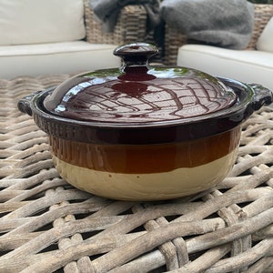 Op de afbeelding: Een bruine en beige keramische ovenschaal met een deksel. De schaal heeft een geglazuurde afwerking en staat op een rieten oppervlak.