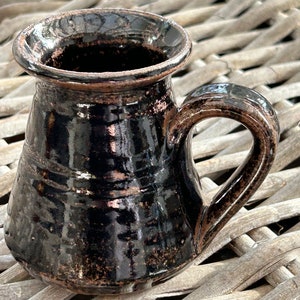 May include: A black ceramic mug with a brown glaze and a handle. The mug has a slightly flared rim and a rounded body.