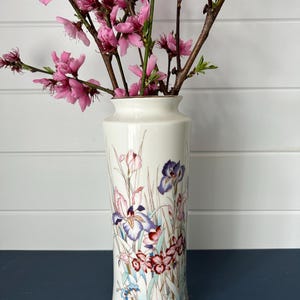 May include: A tall, off-white ceramic vase with a floral design featuring irises and other flowers in shades of purple, pink, and red. The vase holds branches with vibrant pink blossoms. The vase is approximately 10 inches tall.