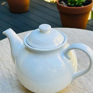 Puede incluir: Una tetera de cerámica blanca con tapa y asa curva. La tetera está sobre una superficie clara y texturizada. Dos macetas de terracota con plantas están en el fondo.