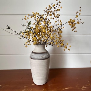 May include: A cream-coloured ceramic vase with a grey and brown band near the top, holding branches of yellow flowers and green leaves. The vase sits on a brown wooden surface.