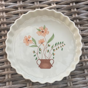 Może przedstawiać: Biała ceramiczna forma do pieczenia z falistym brzegiem. Forma zawiera wzór kwiatowy z pomarańczowymi kwiatami i zielonymi liśćmi w brązowym garnku.