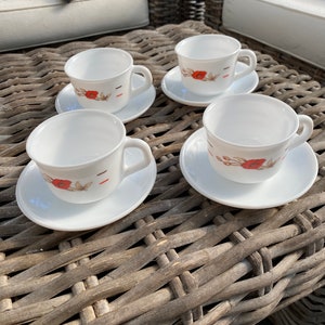 Puede incluir: Cuatro tazas de té de cerámica blanca con diseños florales de amapolas rojas y detalles dorados sobre platillos blancos. Las tazas y los platillos están dispuestos sobre una mesa de mimbre tejido.