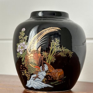 May include: A black ceramic vase with a detailed pheasant design. The vase features a glossy finish and a rounded shape. The pheasant design includes gold, red, and green accents, with floral and foliage elements. The vase is a decorative item.