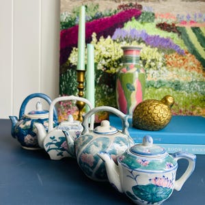 May include: Several small, decorative teapots with floral patterns in blue, white, and pink. The teapots are displayed on a blue surface, with a painting and other ornaments in the background. The teapots vary in size and shape, showcasing a vintage aesthetic.
