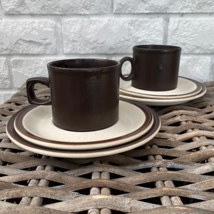 May include: Two brown ceramic coffee mugs with white saucers. The mugs have a brown glaze and a white band around the top. The saucers have a white center with a brown band around the edge.