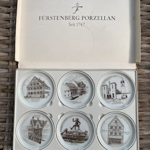 May include: A set of six white porcelain coasters featuring black and white illustrations of buildings and landmarks in Germany. The coasters are presented in a white box with the text "Fürstenberg Porzellan Seit 1747".