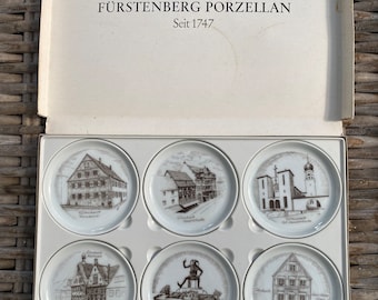 May include: A set of six white porcelain coasters with black and white illustrations of buildings and landmarks in Germany. The coasters are in a white box with the text "Fürstenberg Porzellan Seit 1747".