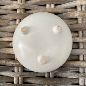 May include: A white ceramic bowl with three small feet on the bottom. The bowl is sitting on a woven wicker surface.