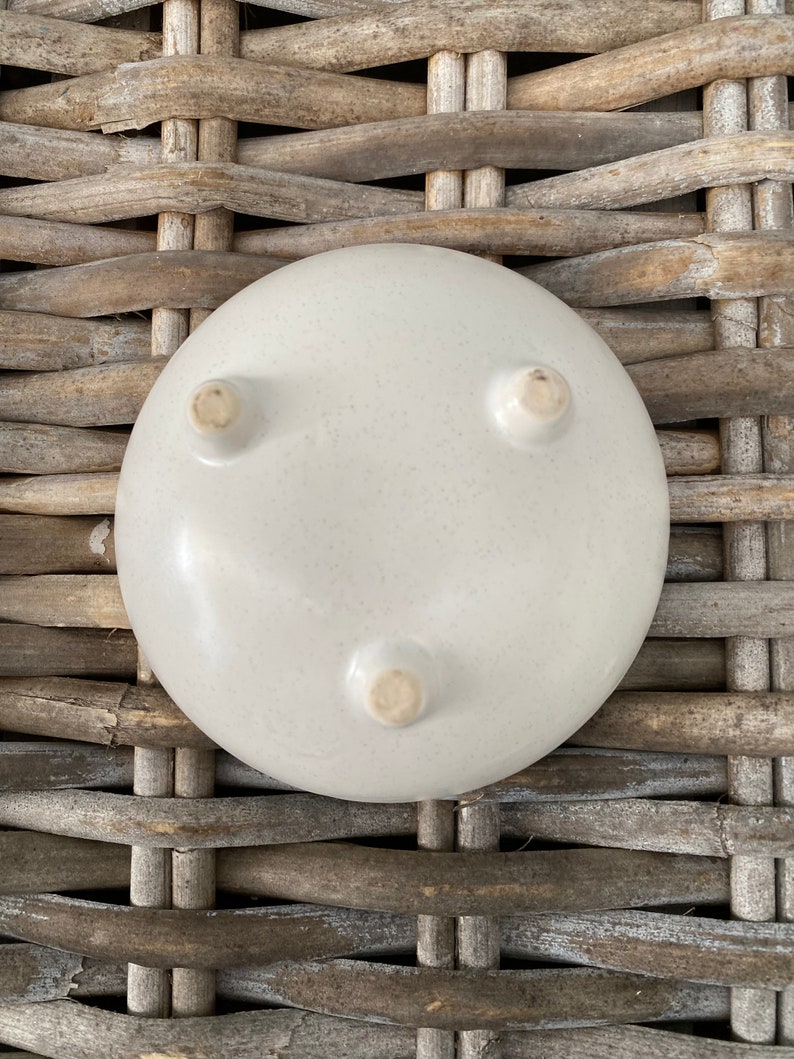 May include: A white ceramic bowl with three small feet on the bottom. The bowl is sitting on a woven wicker surface.