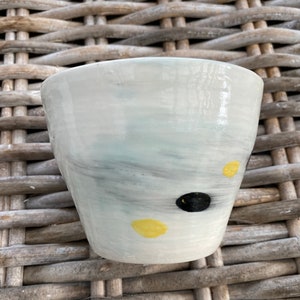 May include: A white ceramic cup with a light blue glaze and black and yellow abstract paint splatters.