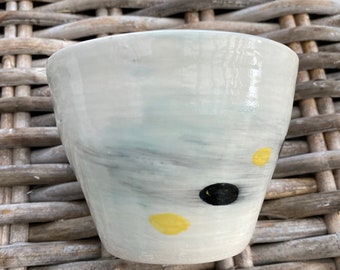 Może przedstawiać: Biały ceramiczny kubek z jasnoniebieską glazurą i czarnymi i żółtymi abstrakcyjnymi plamami farby.