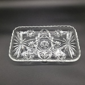 May include: A clear, rectangular glass dish with a decorative starburst and star pattern. The dish has a scalloped edge and is designed with a central starburst and radiating lines. The glass is transparent, allowing light to pass through.