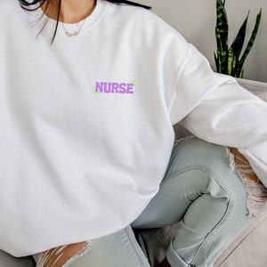 May include: White sweatshirt with the word "NURSE" printed in purple on the front. The sweatshirt is being worn by a person sitting on a couch.