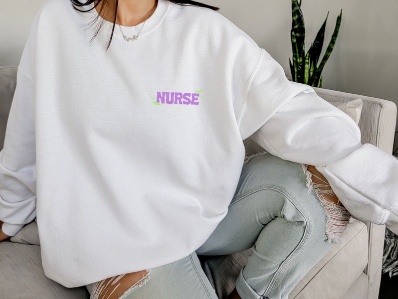 May include: White sweatshirt with the word "NURSE" printed in purple on the front. The sweatshirt is being worn by a person sitting on a couch.