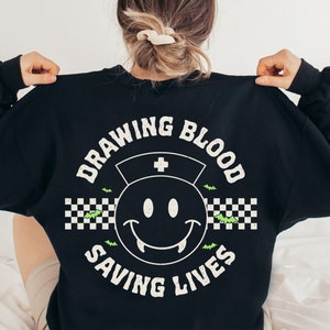 May include: Black sweatshirt with a white graphic print of a smiley face with fangs, a nurse's hat, and the text "Drawing Blood Saving Lives" in a checkered pattern with bats.