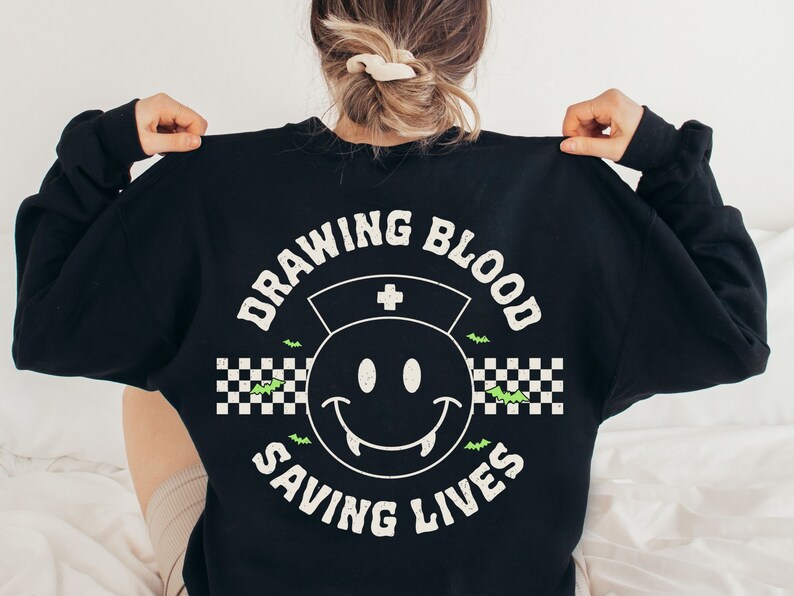 May include: Black sweatshirt with a white graphic print of a smiley face with fangs, a nurse's hat, and the text "Drawing Blood Saving Lives" in a checkered pattern with bats.