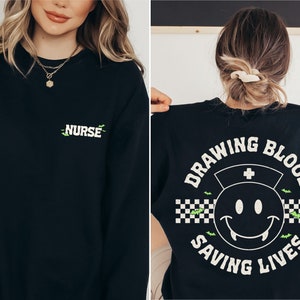 May include: Black sweatshirt with a green and white graphic on the back that says "Drawing Blood Saving Lives" with a smiley face with fangs and a nurse's hat. The front of the sweatshirt has the word "Nurse" in green and white.