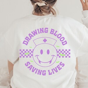 May include: White sweatshirt with a purple graphic design that says "Drawing Blood Saving Lives" with a purple smiley face with fangs and a nurse's hat.