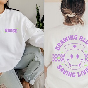 May include: White sweatshirt with a purple "NURSE" text on the front and a purple graphic on the back that says "DRAWING BLOOD SAVING LIVES" with a smiley face wearing a nurse's hat and a checkered pattern.