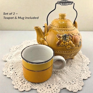 Peut inclure: Un ensemble théière et tasse jaune moutarde. La théière a une anse en fil de fer noir et est décorée de motifs floraux et de fruits en relief. La tasse est jaune avec un intérieur blanc et repose sur un napperon crème. Le texte sur l'image indique : "Set de 2 - Théière et tasse inclus."