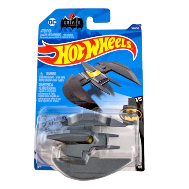 K&ouml;nnte beinhalten: Ein Hot Wheels Spielzeugauto des Batwing aus der Batman-Zeichentrickserie. Der Batwing ist dunkelgrau mit gelben Akzenten, verpackt auf einer blau-roten Karte. Die Verpackung enth&auml;lt die Logos von Hot Wheels und DC Comics.