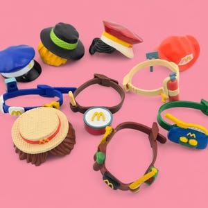 May include: A collection of colorful toy hats and wristbands, including a blue cap, a black hat with a green brim, and a red hat. The wristbands feature various designs and colors, set against a pink background.