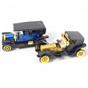 Può includere: Due auto d'epoca in miniatura. Una è blu con tetto nero e dettagli dorati, l'altra è nera e dorata. Entrambe hanno ruote gialle e interni dettagliati. Le auto sono su sfondo bianco.