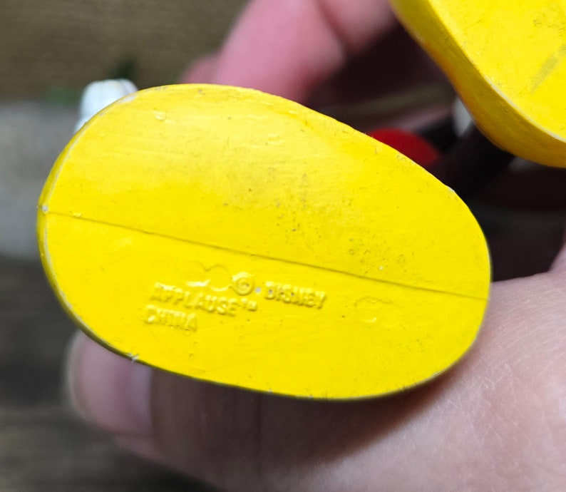 May include: Yellow plastic bottom of a toy figure with the text "Applause" and "Disney" and "China" printed on it.