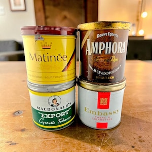 Puede incluir: Cuatro latas de tabaco vintage apiladas. Las latas son amarillas, marrones, blancas y verdes, con varias etiquetas y textos, incluyendo "Matinée", "Amphora", "MacDonald's Export" y "Embassy". Las latas tienen tapas metálicas.