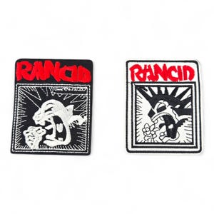 Peut inclure: Deux écussons brodés avec le mot "RANCID" en rouge. L'un présente une panthère blanche rugissante sur fond noir. L'autre affiche un crâne noir et blanc avec des fleurs.