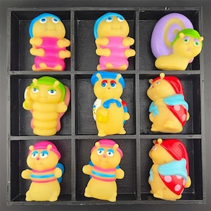 Può includere: Una collezione di nove piccole statuette gialle in stile cartone animato disposte in un vassoio a griglia nero. Le figure presentano vari cappelli, abiti e accessori colorati, tra cui gusci di lumaca, motivi a coccinella e abbigliamento a righe.
