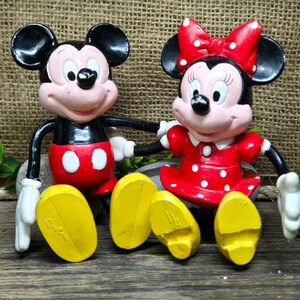 May include: A pair of vintage Mickey Mouse and Minnie Mouse figurines. Mickey is wearing red shorts with white polka dots and yellow shoes. Minnie is wearing a red dress with white polka dots and yellow shoes. Both figurines have black eyes and red noses.