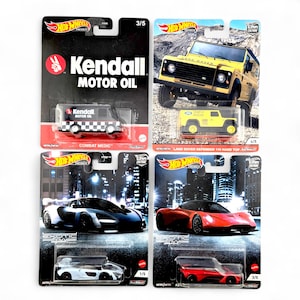 Op de afbeelding: Vier Hot Wheels modelauto's op hun originele verpakking. De collectie bevat een zwart-witte Kendall Motor Oil Combat Medic truck, een gele Land Rover Defender en twee Exotic Envy sportwagens in zwart en rood. De verpakking toont stadsgezichten.