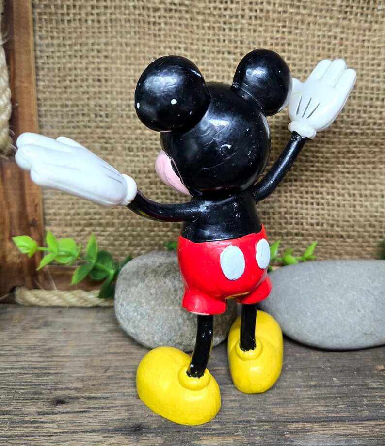 May include: A black and red plastic Mickey Mouse figurine with white gloves and yellow shoes. The figurine is standing with its arms raised and its head tilted back.