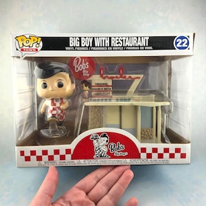 May include: A Pop! Town vinyl figure set featuring Big Boy with a restaurant diorama. The figure is of a smiling boy in a checkered outfit. The restaurant has a diner-style design with the Bob's Big Boy logo and the words "Original Double Deck Hamburger".