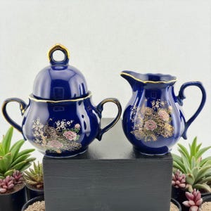 Peut inclure: Un sucrier en céramique bleu marine avec un couvercle et une crémière. Les deux pièces ont des bordures dorées et des motifs floraux avec des accents roses et dorés. Le sucrier a une poignée de chaque côté et une boucle sur le couvercle.
