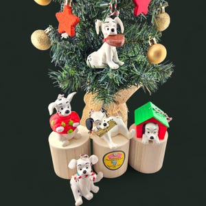 Peut inclure: Un sapin de Noël orné de décorations sur le thème des Dalmatiens. Les ornements représentent des figurines de chiens dans diverses poses, certains tenant des objets. L'arbre est également décoré d'étoiles rouges et de boules dorées.