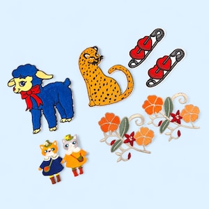 以下が含まれることがあります： 様々なデザインの刺繍パッチのコレクション。赤いリボンが付いた青い子羊、黒い斑点のある黄色のチーター、2つの赤いハート型のピン、2つの猫のフィギュア、オレンジ色の花柄のデザインが含まれています。