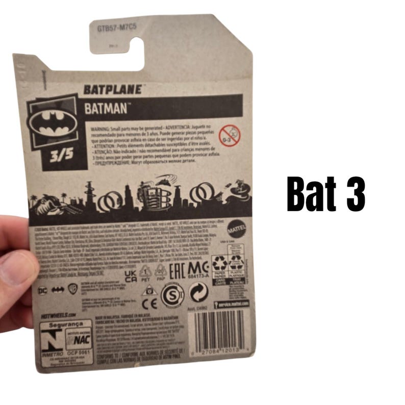 K&ouml;nnte beinhalten: Kartonverpackung f&uuml;r ein Hot Wheels Batplane-Spielzeug. Die Verpackung zeigt das Batman-Logo, den Text "BATPLANE BATMAN" und die Nummer "3/5". Die R&uuml;ckseite der Verpackung enth&auml;lt Sicherheitshinweise und einen Barcode.