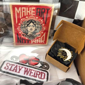 Peut inclure: Un assortiment d'écussons et une épingle. L'écusson rouge porte l'inscription "MAKE ART NOT WAR" avec un visage de femme au centre. Une épingle dans une boîte présente un motif soleil et lune. L'écusson "STAY WEIRD" est également visible.