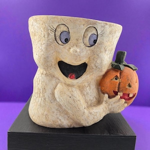 Peut inclure: Une figurine de fantôme en céramique sur le thème d'Halloween, avec un visage souriant, tenant une lanterne citrouille orange. Le fantôme a de grands yeux bleus et une finition blanc cassé craquelée. La lanterne citrouille a un visage sculpté et une tige verte.