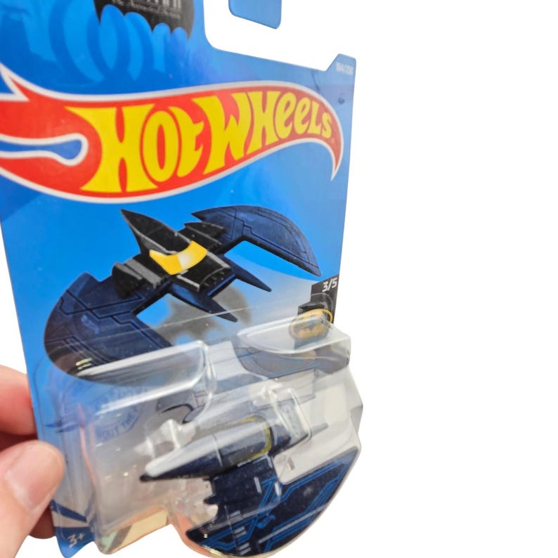 K&ouml;nnte beinhalten: Ein Hot Wheels Batman Batwing Spielzeugauto aus Druckguss in Blau und Gelb. Das Spielzeug ist in einer blauen und transparenten Blisterverpackung mit dem Hot Wheels-Logo und Batman-Branding verpackt.
