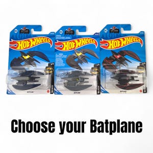 K&ouml;nnte beinhalten: Drei Hot Wheels Batplane Spielzeugautos in ihrer Originalverpackung. Die Batplanes sind schwarz mit gelben oder roten Akzenten. Die Verpackung ist blau mit den Hot Wheels- und Batman-Logos. Der Text auf dem Bild lautet "W&auml;hle dein Batplane."
