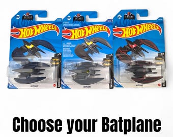 Könnte beinhalten: Drei Hot Wheels Batplane Spielzeugautos in ihrer Originalverpackung. Die Batplanes sind schwarz mit gelben oder roten Akzenten. Die Verpackung ist blau mit den Hot Wheels- und Batman-Logos. Der Text auf dem Bild lautet "Wähle dein Batplane."