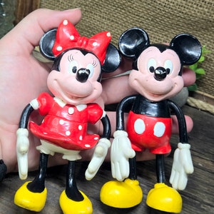 May include: Two plastic figurines of Mickey Mouse and Minnie Mouse. Mickey Mouse is wearing red shorts and black shoes. Minnie Mouse is wearing a red dress with white polka dots and black shoes. Both figurines have black eyes and red cheeks.