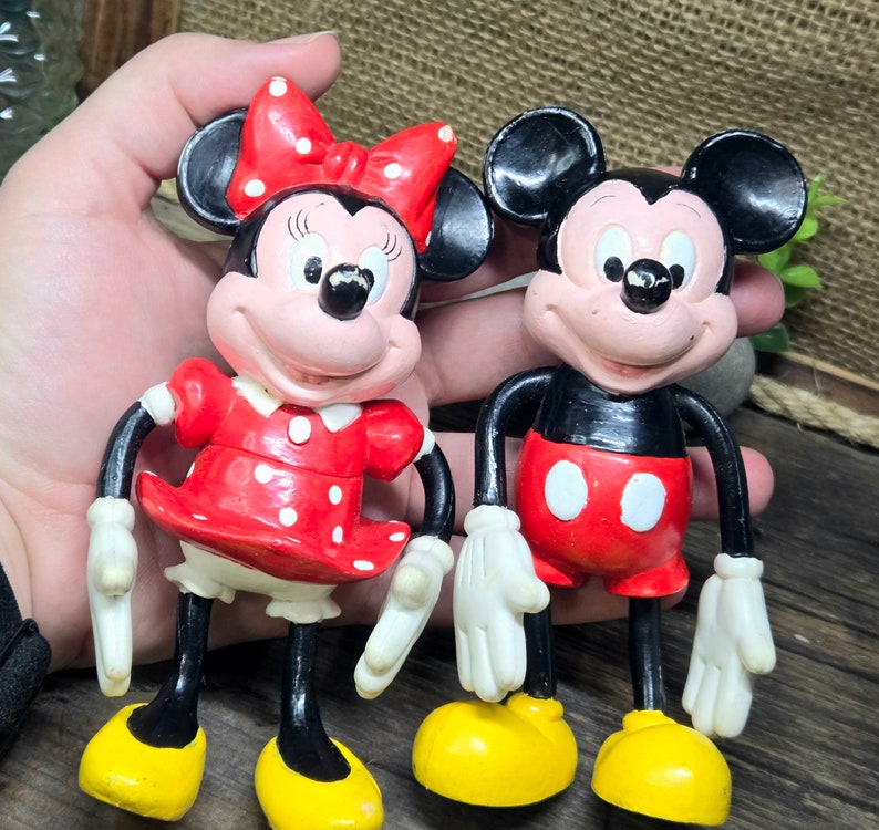 May include: Two plastic figurines of Mickey Mouse and Minnie Mouse. Mickey Mouse is wearing red shorts and black shoes. Minnie Mouse is wearing a red dress with white polka dots and black shoes. Both figurines have black eyes and red cheeks.