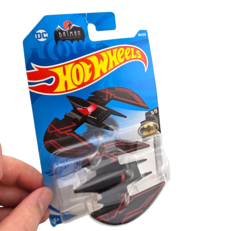 K&ouml;nnte beinhalten: Ein schwarzes Hot Wheels Batwing Spielzeugauto mit roten Akzentlinien, verpackt auf einer blauen Karte mit den Hot Wheels und Batman Logos. Das Spielzeugauto befindet sich in einer transparenten Blisterverpackung.