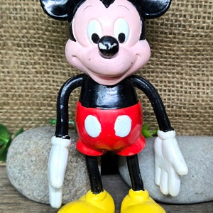 May include: A vintage plastic Mickey Mouse figurine with black ears, a black body, red shorts with white circles, yellow shoes, and a smiling face.